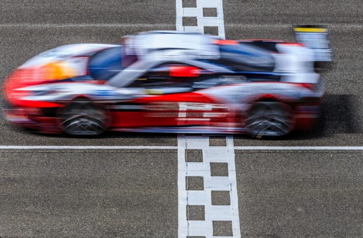 A red and white race car speeding across a finish line with motion blur, captured from above on a racetrack.