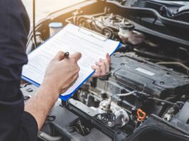 A mechanic going over a maintenance checklist on a clipboard in front of a car with the hood popped open.