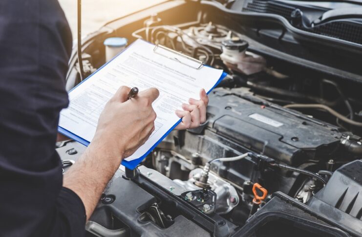 A mechanic going over a maintenance checklist on a clipboard in front of a car with the hood popped open.