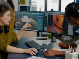 Two coworkers review 3D design models on dual monitors, one pointing at the screen during a desk discussion.