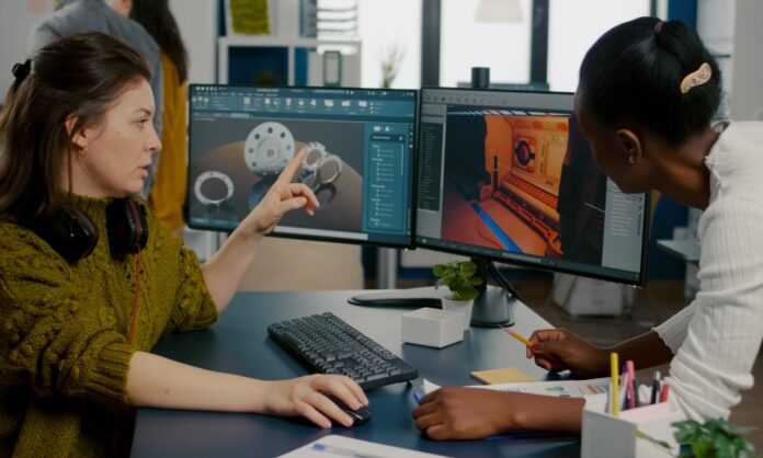 Two coworkers review 3D design models on dual monitors, one pointing at the screen during a desk discussion.