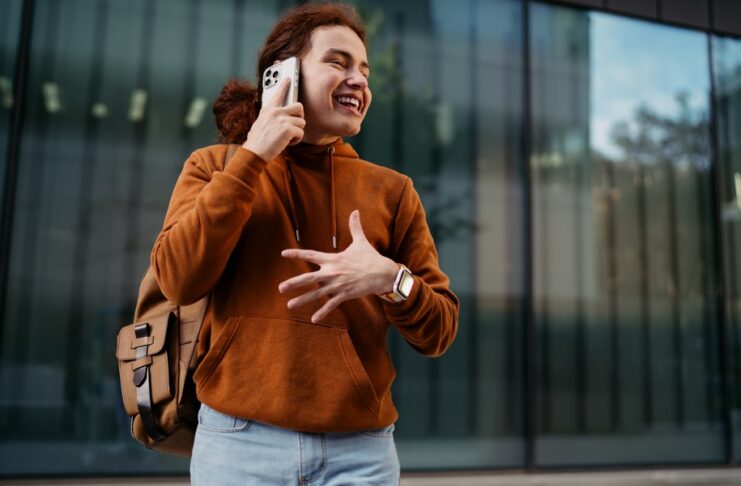 Smiling person with a shoulder bag holds a smartphone to their ear in front of tall reflective windows.