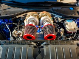 A close-up view of a car engine bay with black plastic covers, hoses, wiring, and a red battery terminal.