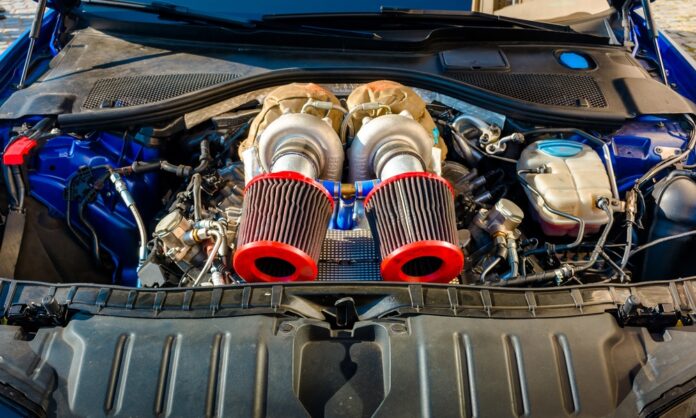 A close-up view of a car engine bay with black plastic covers, hoses, wiring, and a red battery terminal.