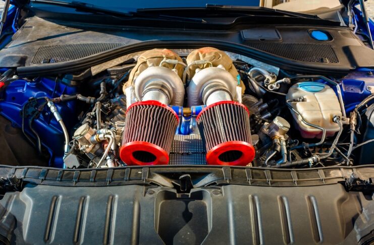 A close-up view of a car engine bay with black plastic covers, hoses, wiring, and a red battery terminal.