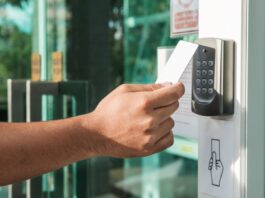 A person holding a white RFID key card to an electronic access control reader at the entrance of a modern office building.