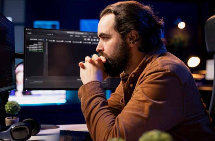 Man focuses on several computer screens at a workstation, with headphones nearby in a dark, blue-lit office.
