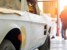 A man is standing in the background while there is a car in the foreground. The car is in a state of disrepair.