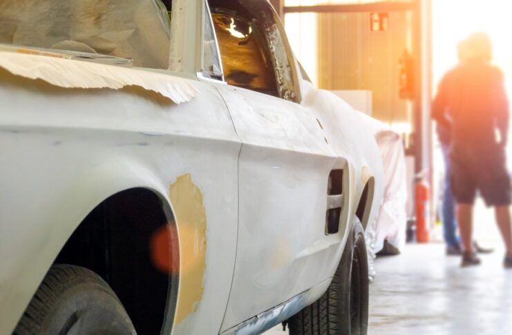 A man is standing in the background while there is a car in the foreground. The car is in a state of disrepair.