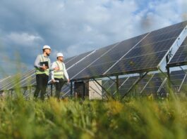 Two engineers in hard hats and safety vests walking through grass beside rows of solar panels under a cloudy sky.