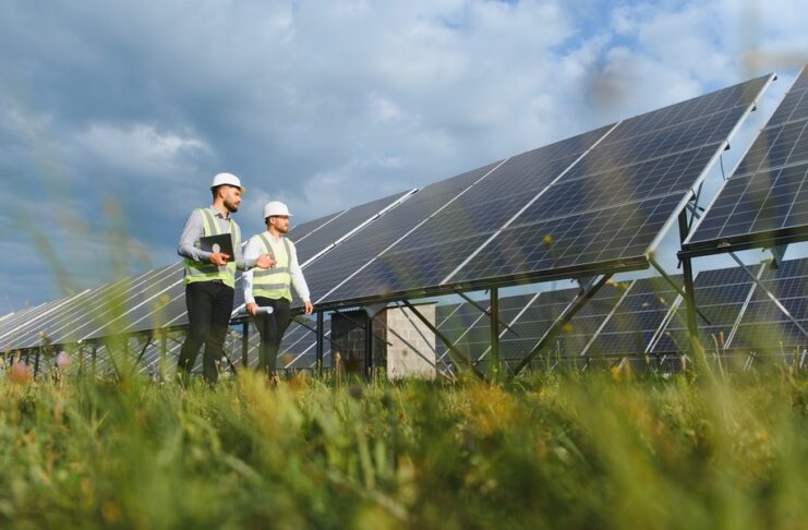 Two engineers in hard hats and safety vests walking through grass beside rows of solar panels under a cloudy sky.