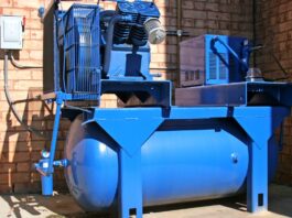Blue industrial air compressor with a large horizontal storage tank installed beside a brick wall and electrical control box.