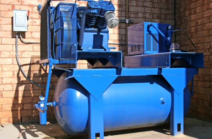 Blue industrial air compressor with a large horizontal storage tank installed beside a brick wall and electrical control box.
