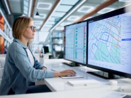 A woman wearing glasses sits at a white desk using a computer with map routes displayed on dual monitors.