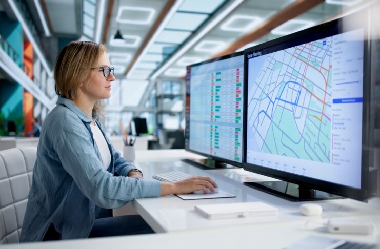 A woman wearing glasses sits at a white desk using a computer with map routes displayed on dual monitors.