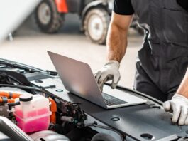 A person uses a laptop beside an open car engine compartment with tools and mechanical parts visible nearby.