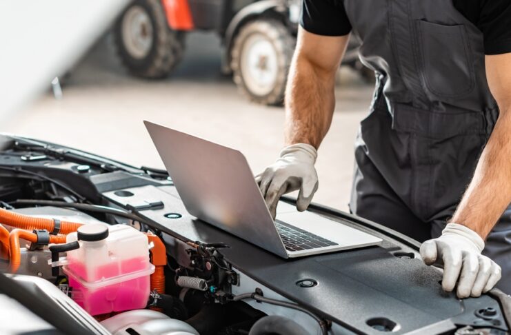 A person uses a laptop beside an open car engine compartment with tools and mechanical parts visible nearby.