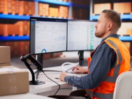 A warehouse worker monitors inventory data on computer screens beside stacked cardboard boxes and a barcode scanner.
