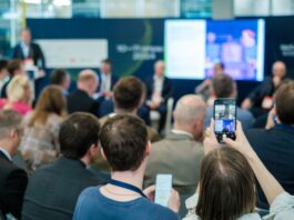An audience seated at a conference while a person holds a smartphone toward speakers and a large screen.