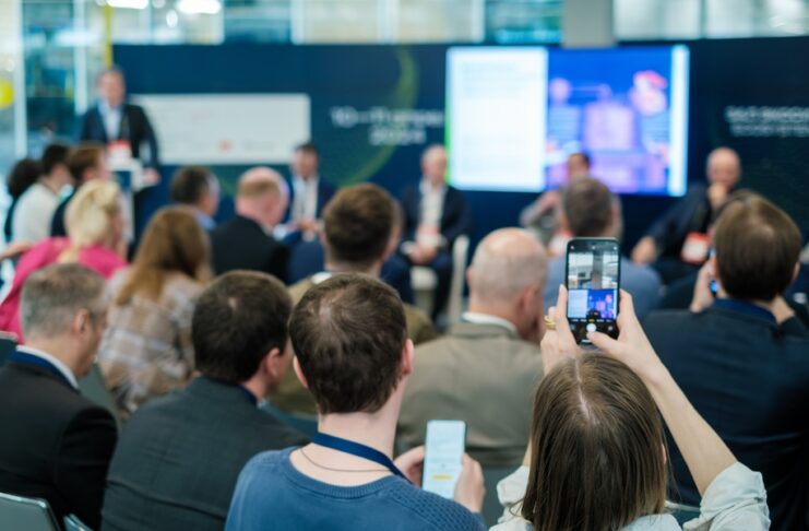 An audience seated at a conference while a person holds a smartphone toward speakers and a large screen.