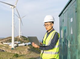 A technician uses a tablet beside an outdoor equipment enclosure with wind turbines operating in the background.
