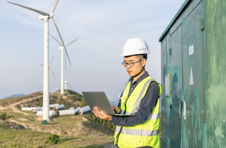 A technician uses a tablet beside an outdoor equipment enclosure with wind turbines operating in the background.