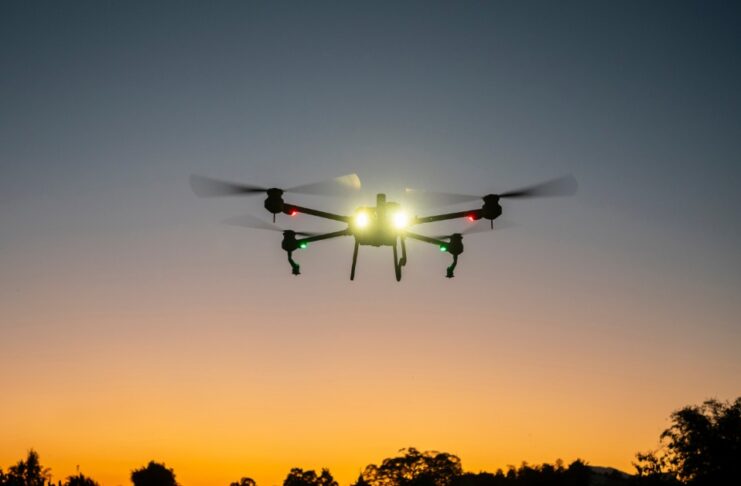 A drone with lights is flying above a field. Trees and an orange and pink sunset are in the background.