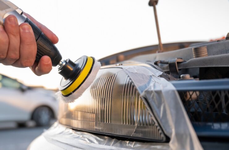 A person using a polishing tool on a car headlight outdoors, with the vehicle's front and pavement nearby.