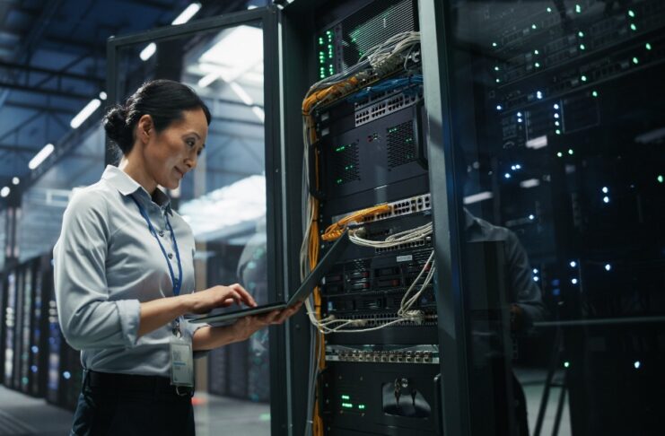 A woman using a laptop beside an open server rack with cables and glowing equipment in a large data center.
