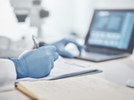 A close-up view shows a scientist writing on a piece of paper with one hand and touching a laptop keyboard with the other.