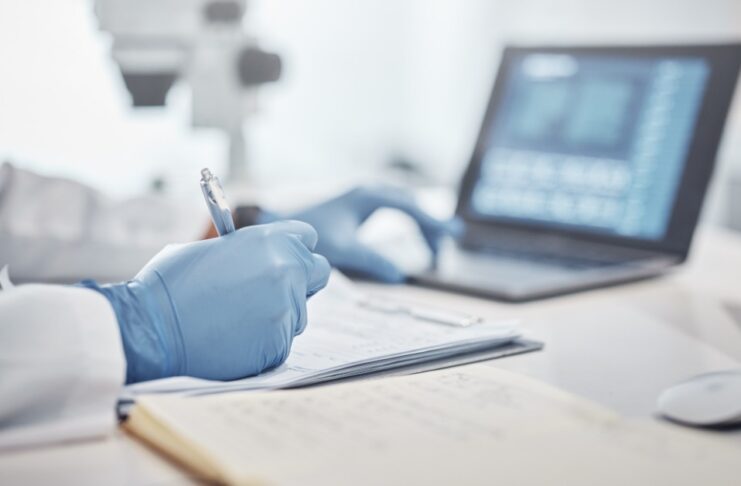 A close-up view shows a scientist writing on a piece of paper with one hand and touching a laptop keyboard with the other.
