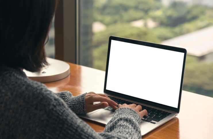 An over-the-shoulder view shows a woman sitting at a desk typing on a laptop keyboard. The desk faces a large window.