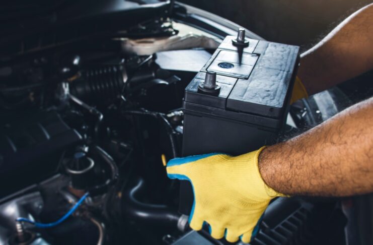 A mechanic wearing yellow-and-green gloves to remove a large black battery from a car in a repair shop.