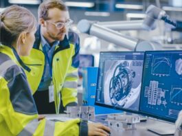 Two engineers wearing high-visibility vests by a computer monitor in factory that shows a 3D mechanical part.