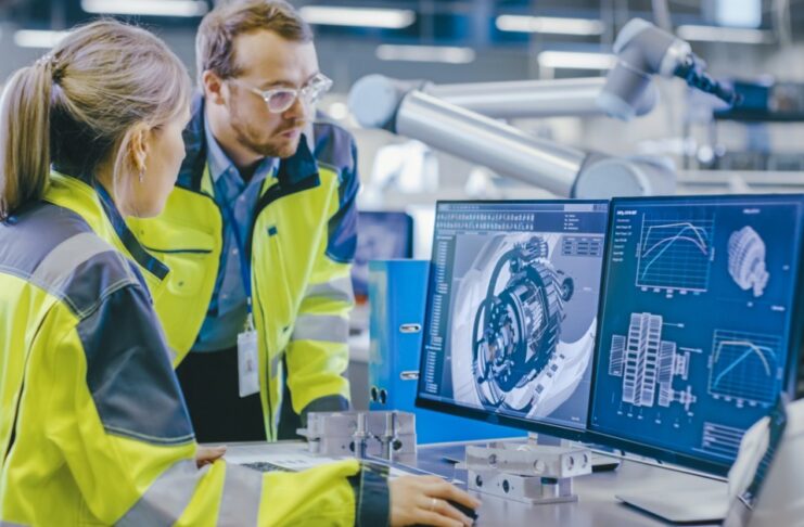 Two engineers wearing high-visibility vests by a computer monitor in factory that shows a 3D mechanical part.