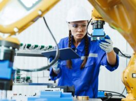 An engineer wearing a hard hat and safety glasses inspecting a yellow robotic arm while holding a clipboard.