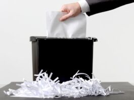 A person wearing a suit jacket places a document into a black paper shredder. Paper strips are scattered across the table.