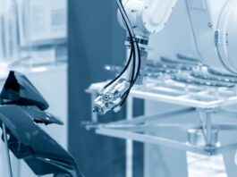 A white robotic arm with a metal spray nozzle pointing toward dark motorcycle body panels in a blue-tinted factory.
