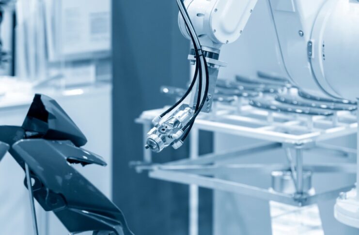 A white robotic arm with a metal spray nozzle pointing toward dark motorcycle body panels in a blue-tinted factory.