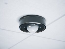 Black circular ceiling-mounted motion sensor with a white dome and blue indicator light installed on white ceiling tiles.